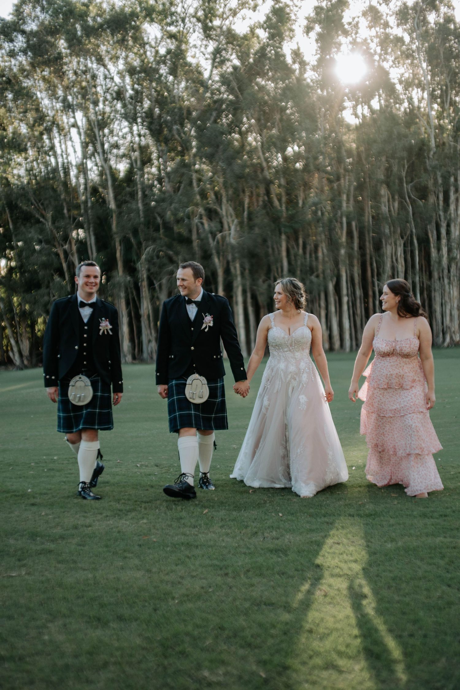 Wedding party in formal attire walks through a sunlit eucalyptus grove casting long shadows on green grass.