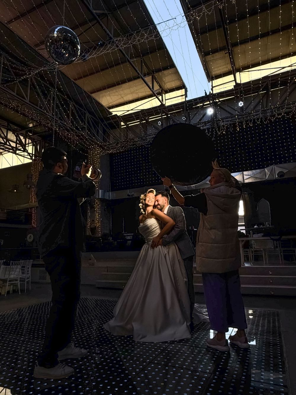 A couple dances together in a dimly lit venue with string lights overhead during what appears to be a wedding reception.