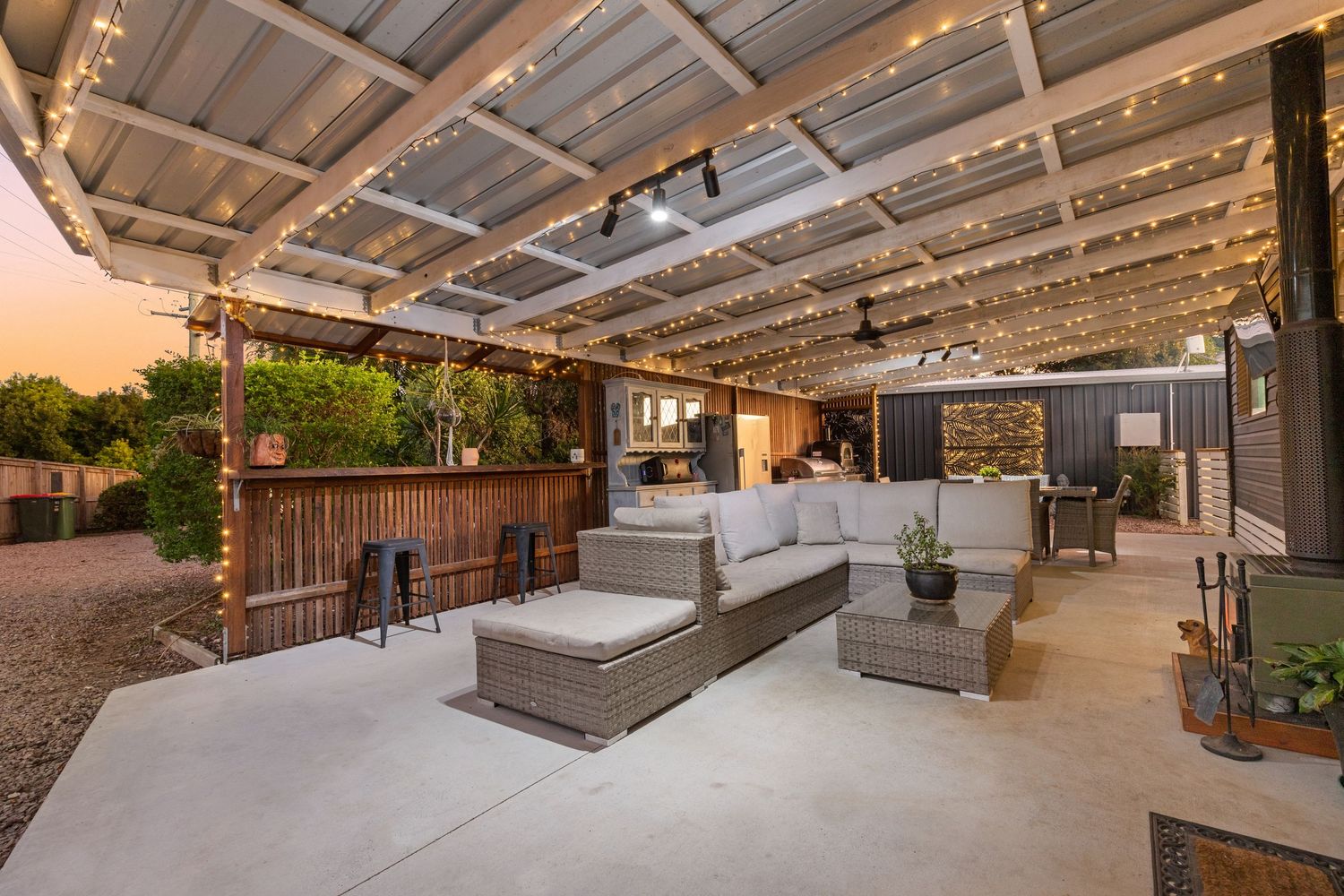 Alfresco entertaining area with string lights, rattan furniture, outdoor kitchen, and pergola roof at dusk.