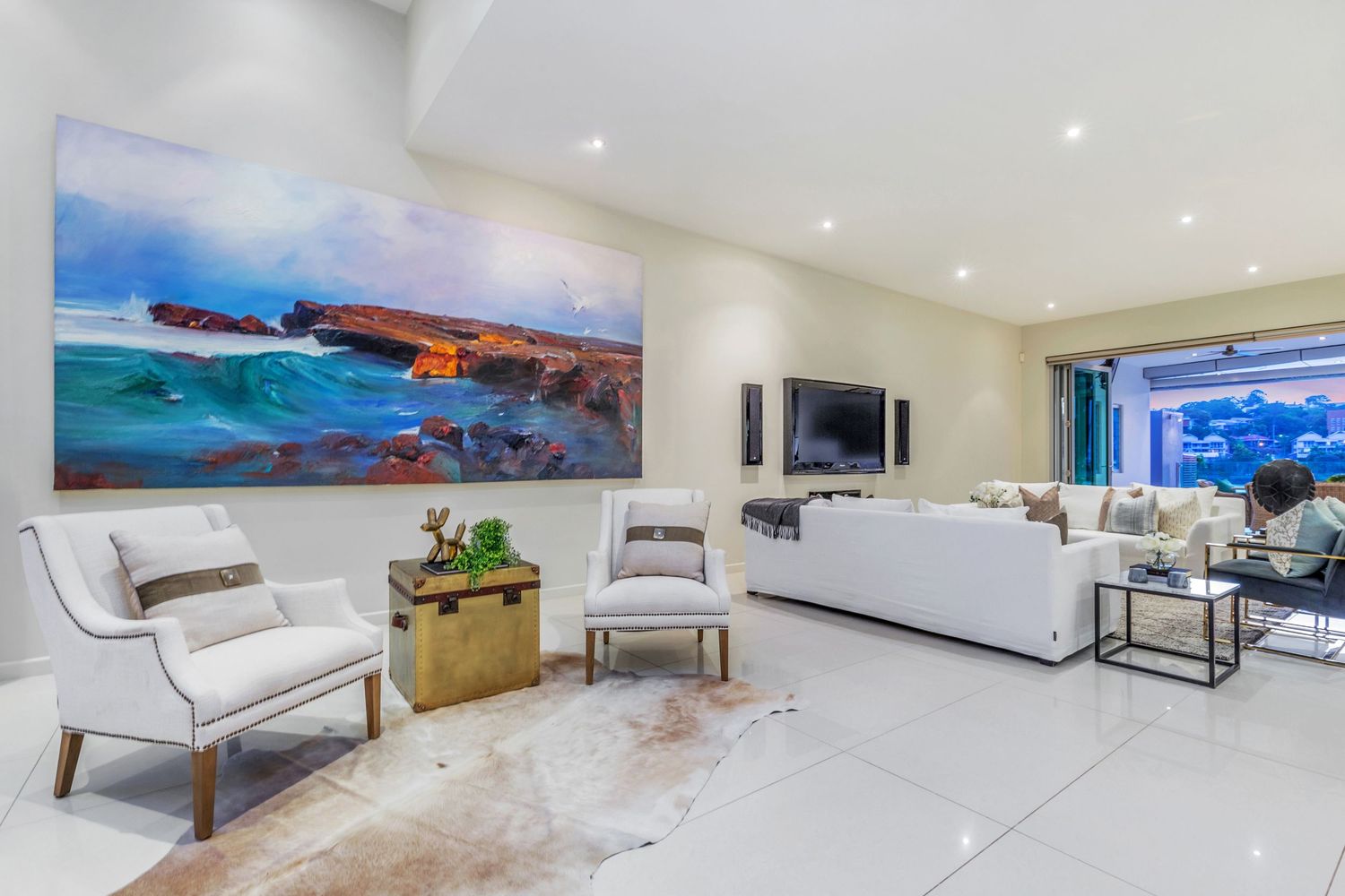 Elegant living room with white sofas, large coastal artwork, marble coffee table, and recessed lighting on white tiles.