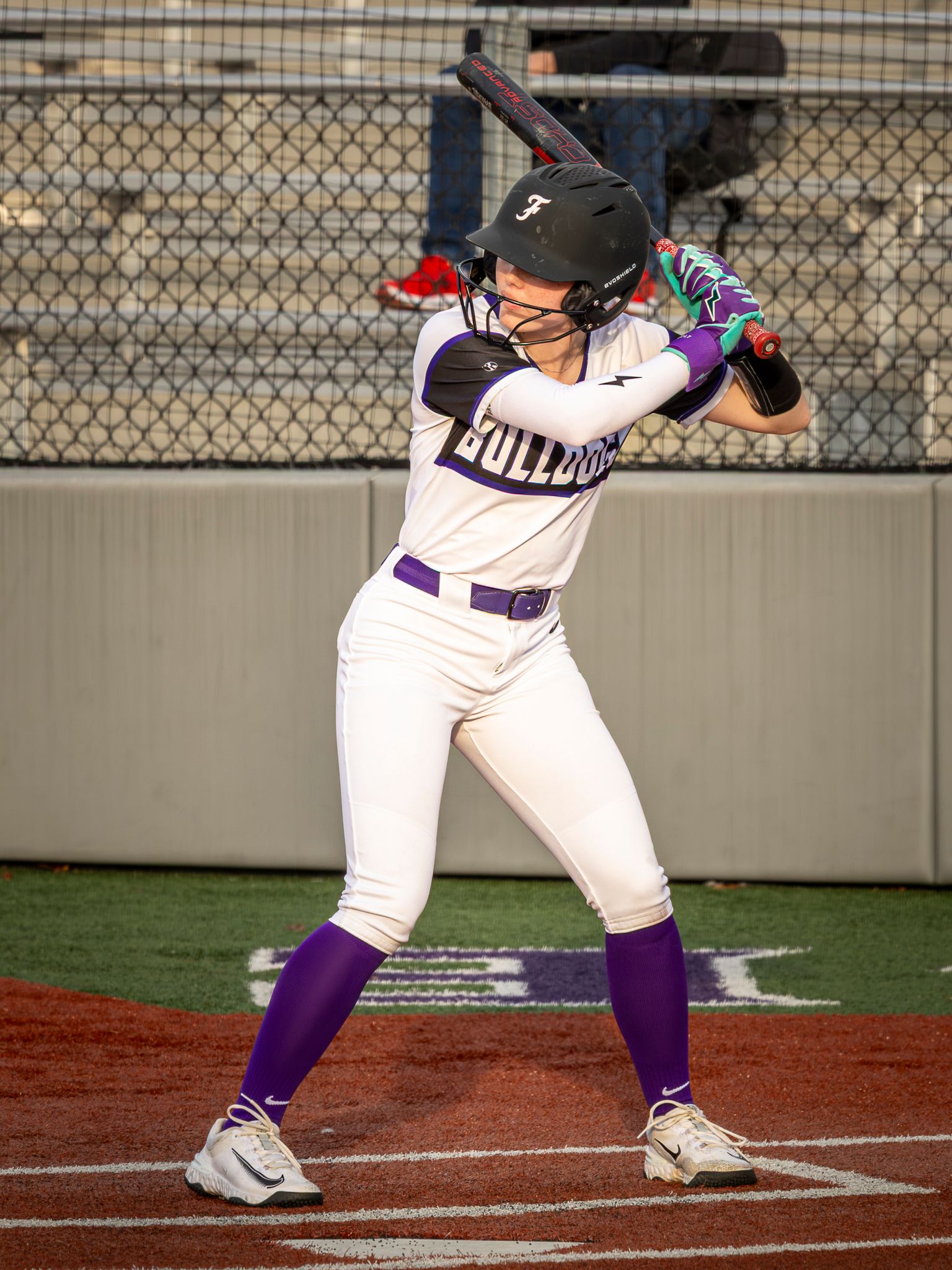 Baseball batter in white and purple uniform demonstrates proper batting stance at home plate during a game.