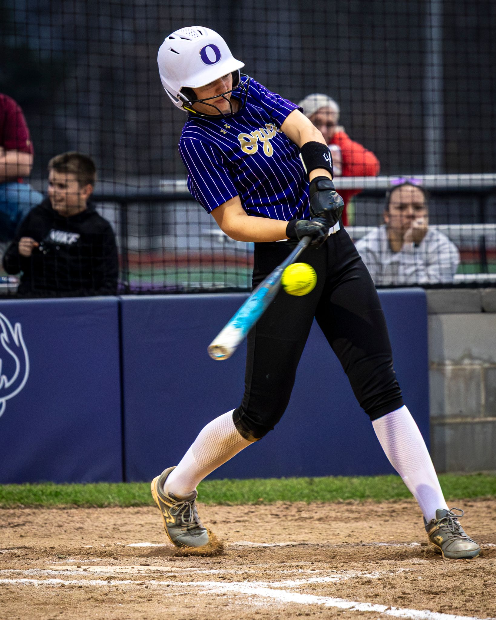 A softball player in a pinstriped uniform swings at a yellow ball during a game, with spectators visible in the background.
