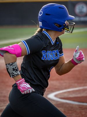 A Rogers softball player wearing pink batting gloves and a blue helmet swings through a pitch at home plate.