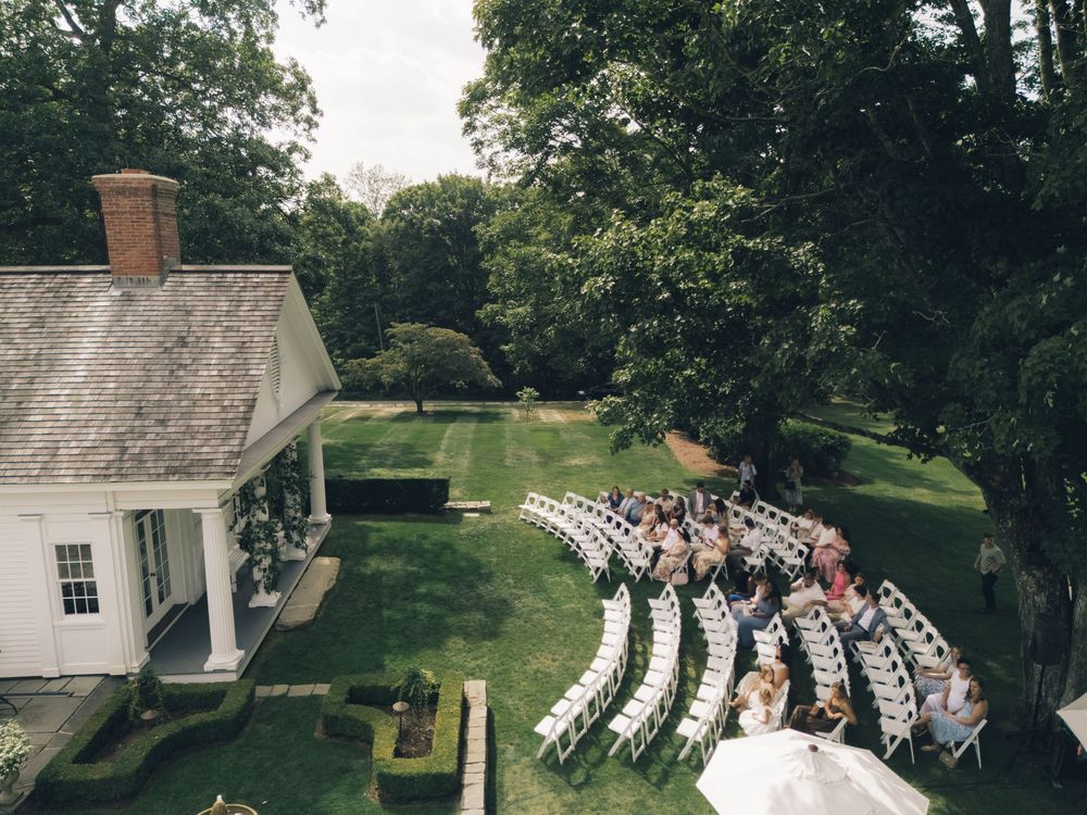 Outdoor wedding ceremony setup with white chairs arranged in curved rows on a lush green lawn surrounded by trees.