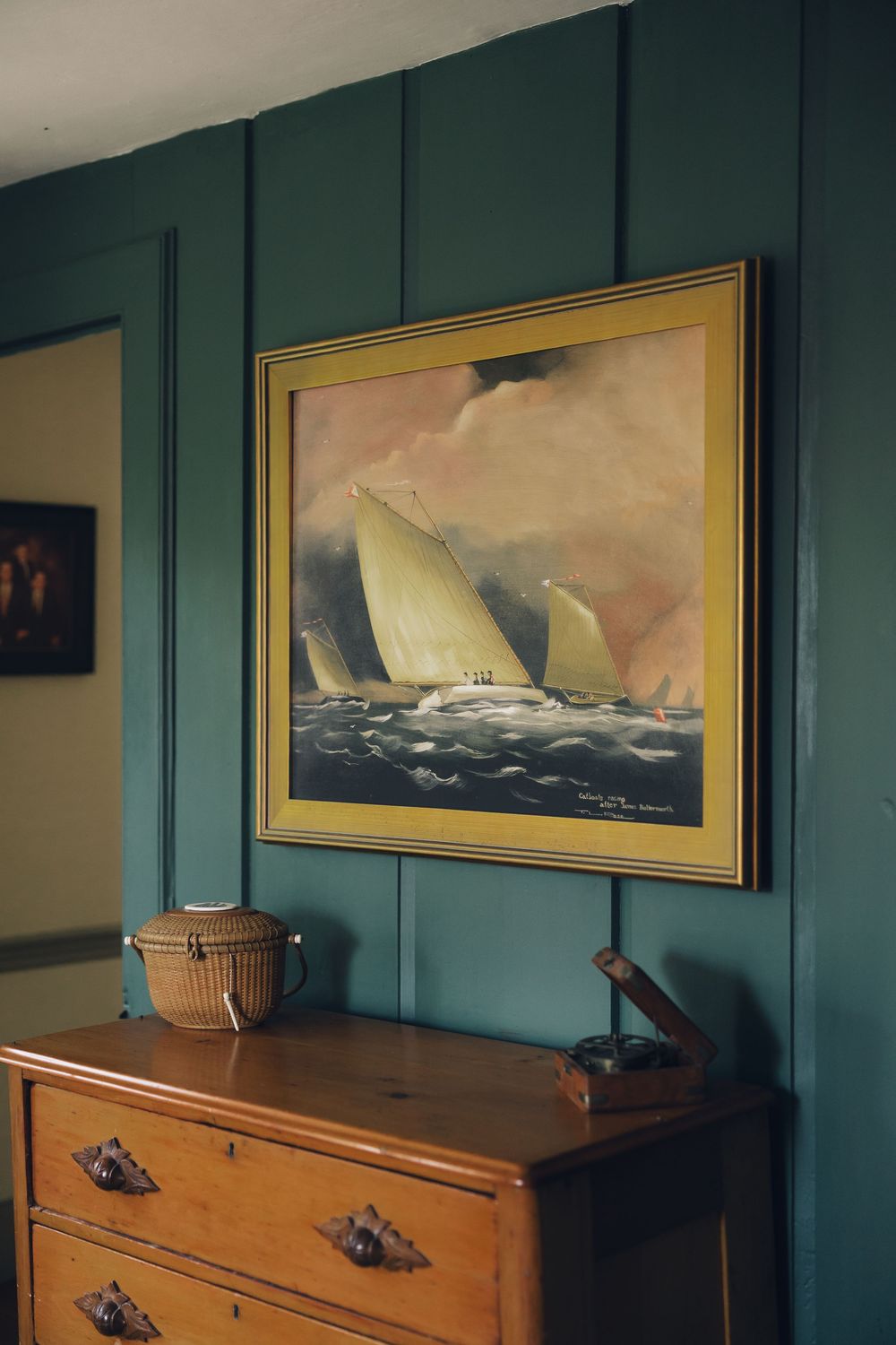 A nautical painting of sailboats at sea hangs above a wooden dresser against a dark green wall.
