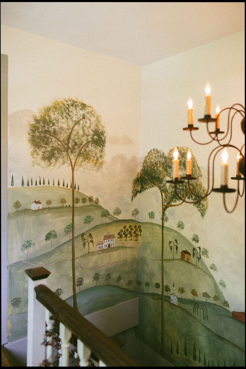 A mural depicting a pastoral countryside landscape with rolling hills and trees, illuminated by a wall-mounted candelabra.