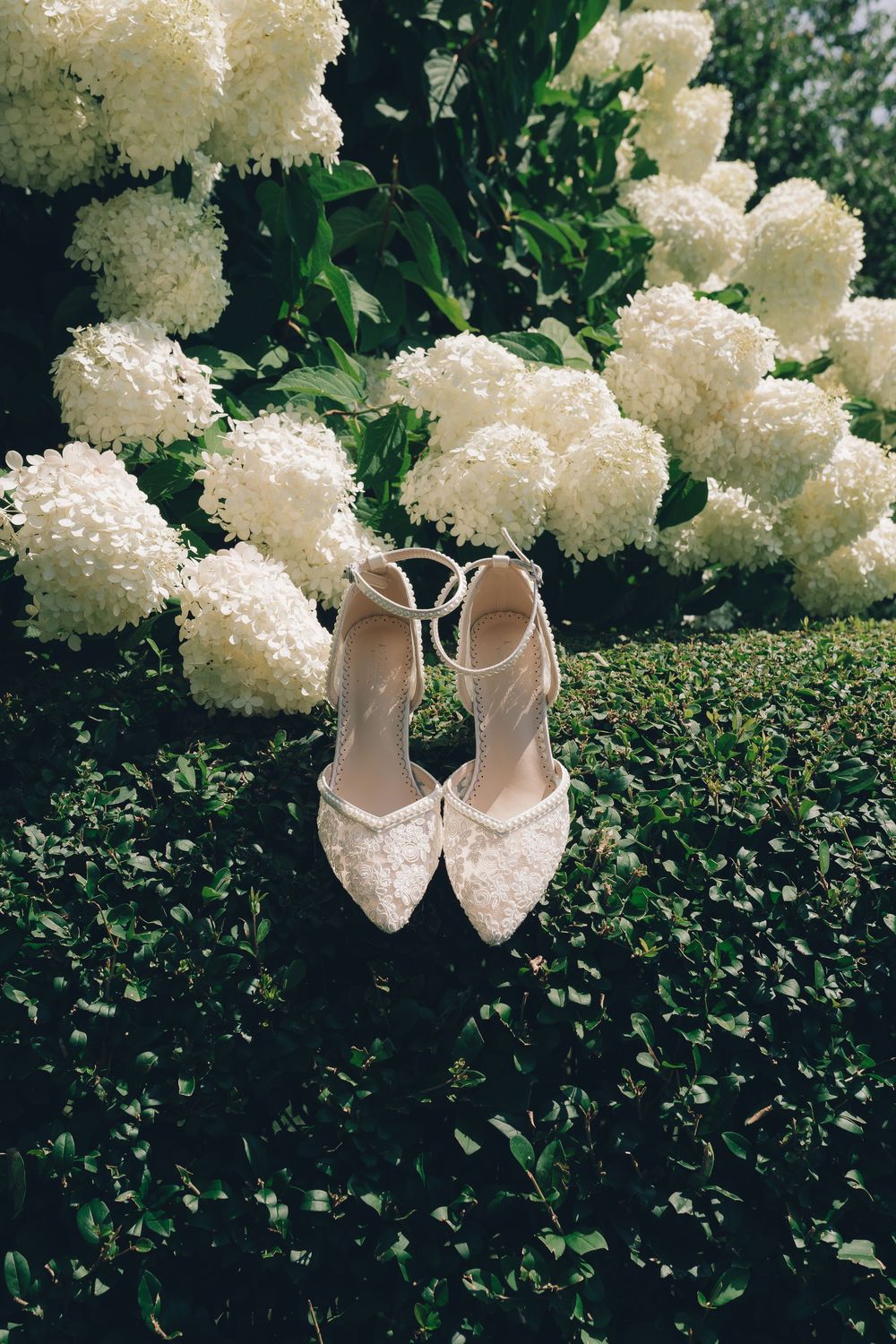 Elegant white pearl-studded bridal shoes displayed among blooming white hydrangea flowers and green foliage.