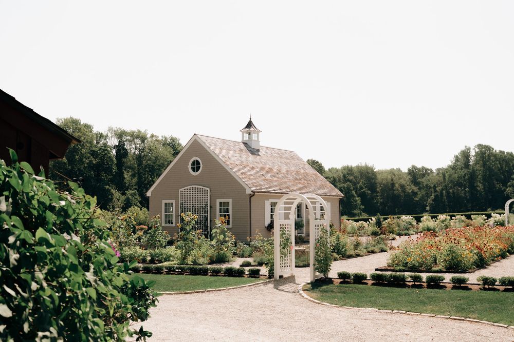 A charming tan chapel with white trim and steeple sits among lush gardens and trees on a gravel path.
