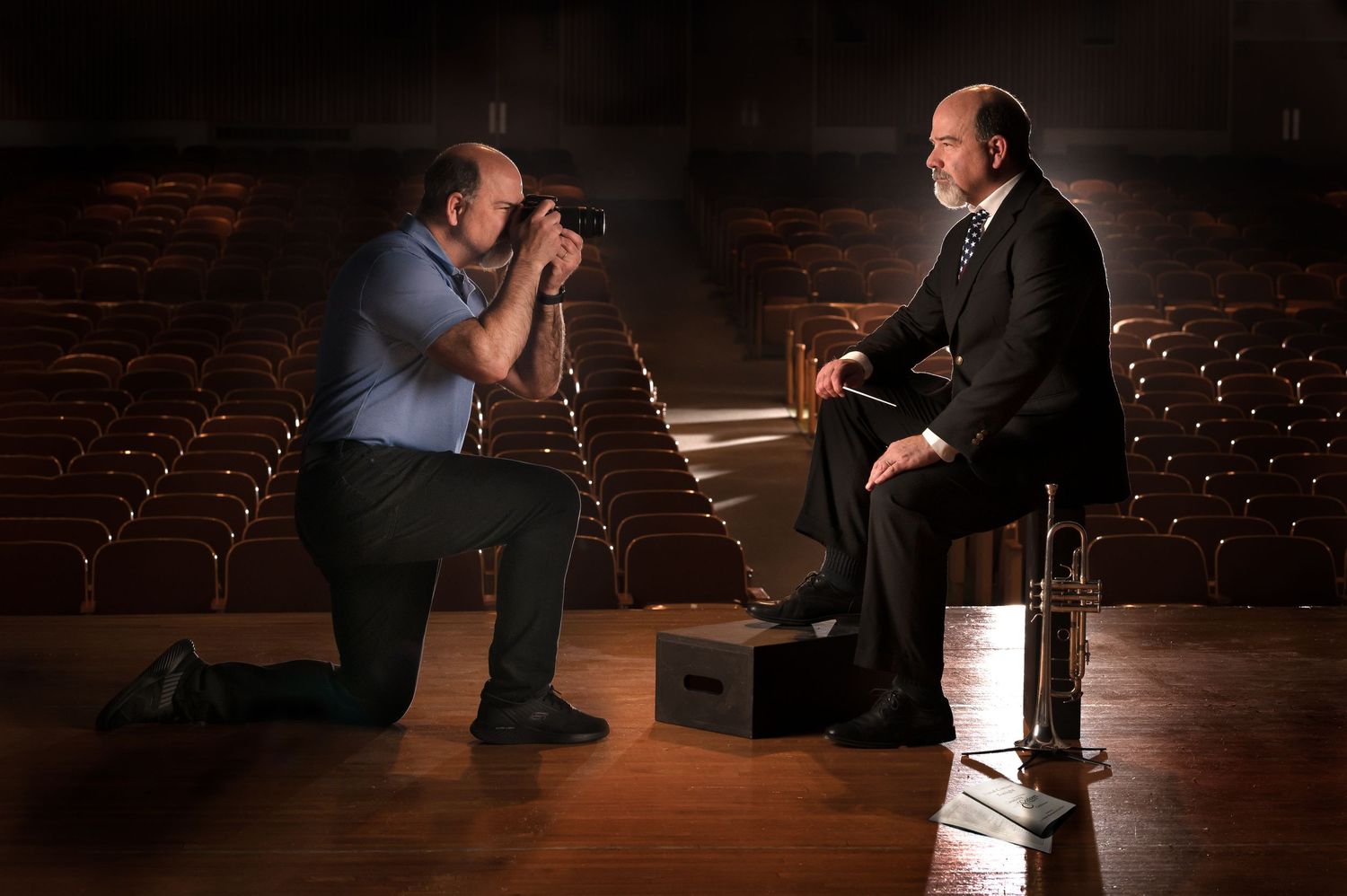 Two figures engage in a dramatic scene on a dimly lit stage with theatrical lighting creating moody shadows.