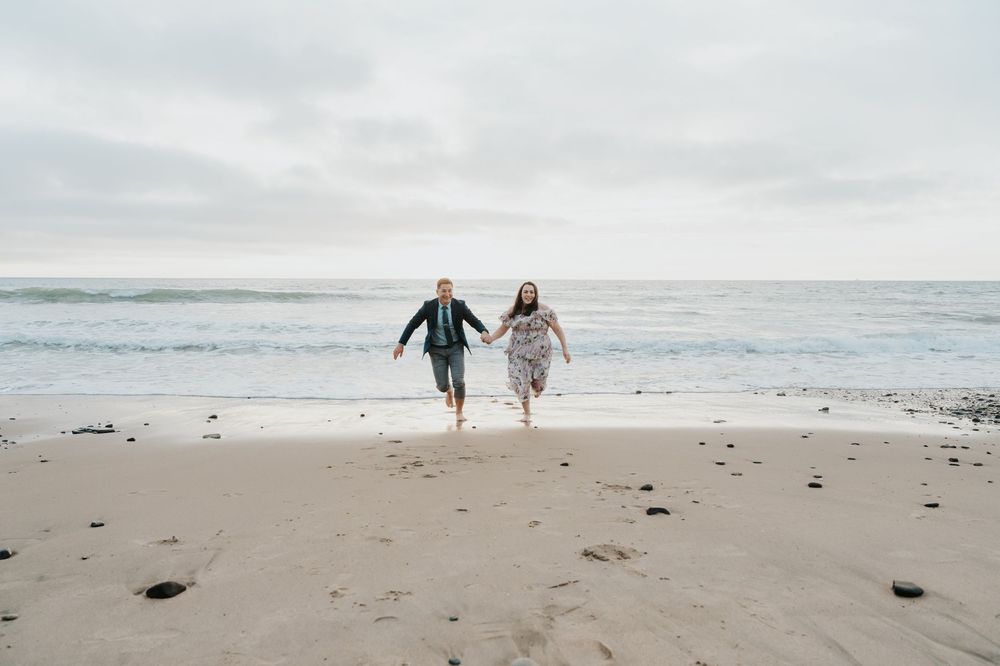 Two people walking hand in hand along a misty beach shoreline with waves crashing in the background.