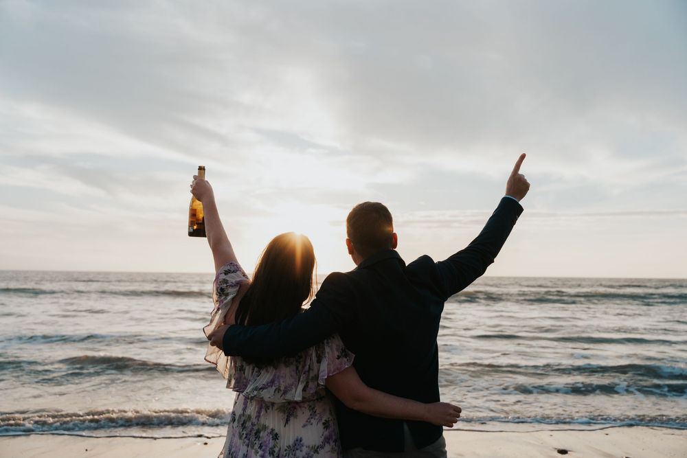 Two people celebrating with raised arms while watching sunset at the beach with one holding a bottle.