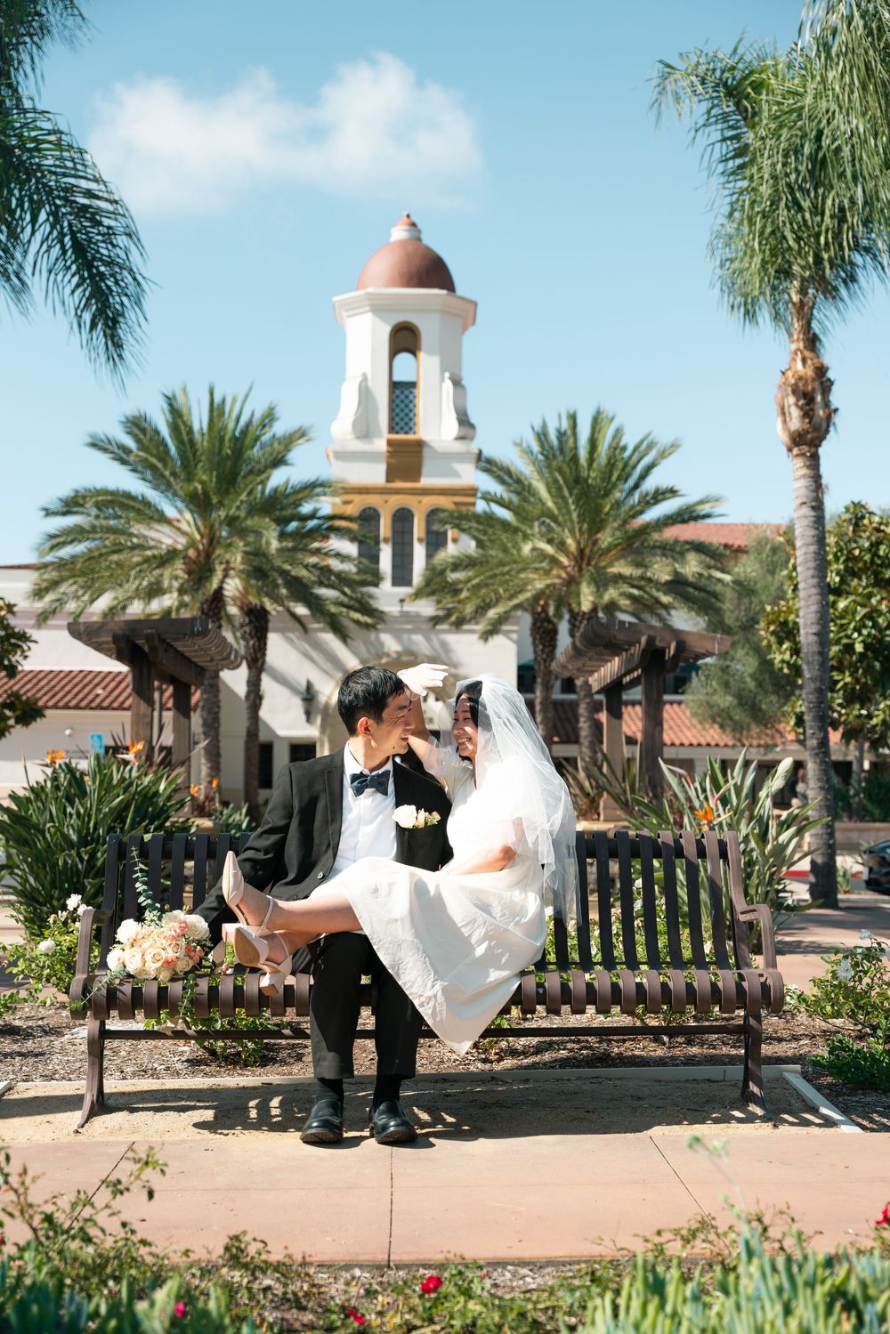 A romantic wedding couple sits on a bench in front of a Spanish-style church tower surrounded by palm trees and gardens.