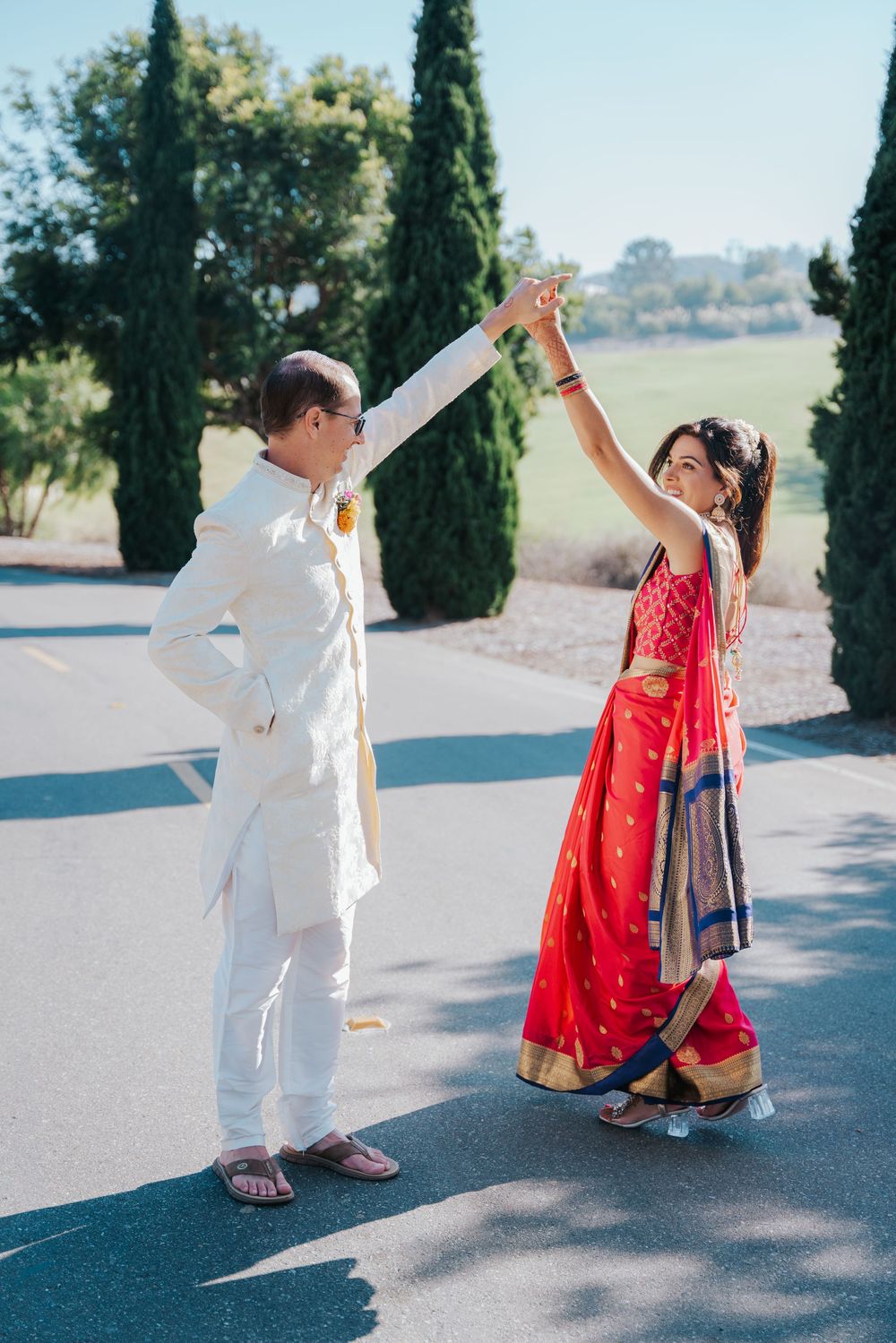 A couple in traditional Indian attire dances together in bright sunlight on a paved outdoor area.