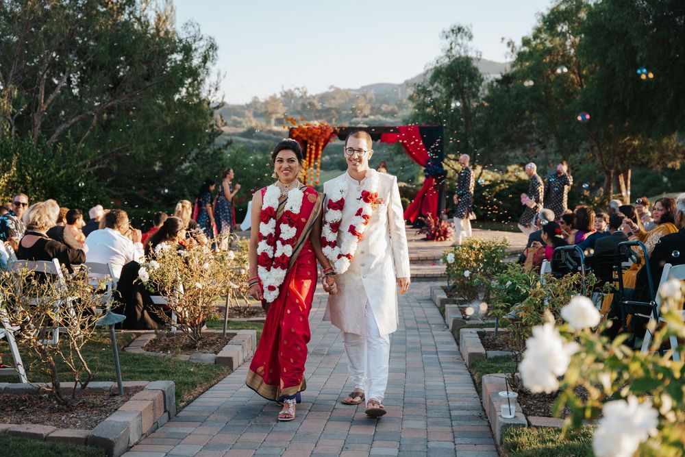 Couple walks down aisle in traditional Indian wedding attire at outdoor ceremony with guests and floral arch.
