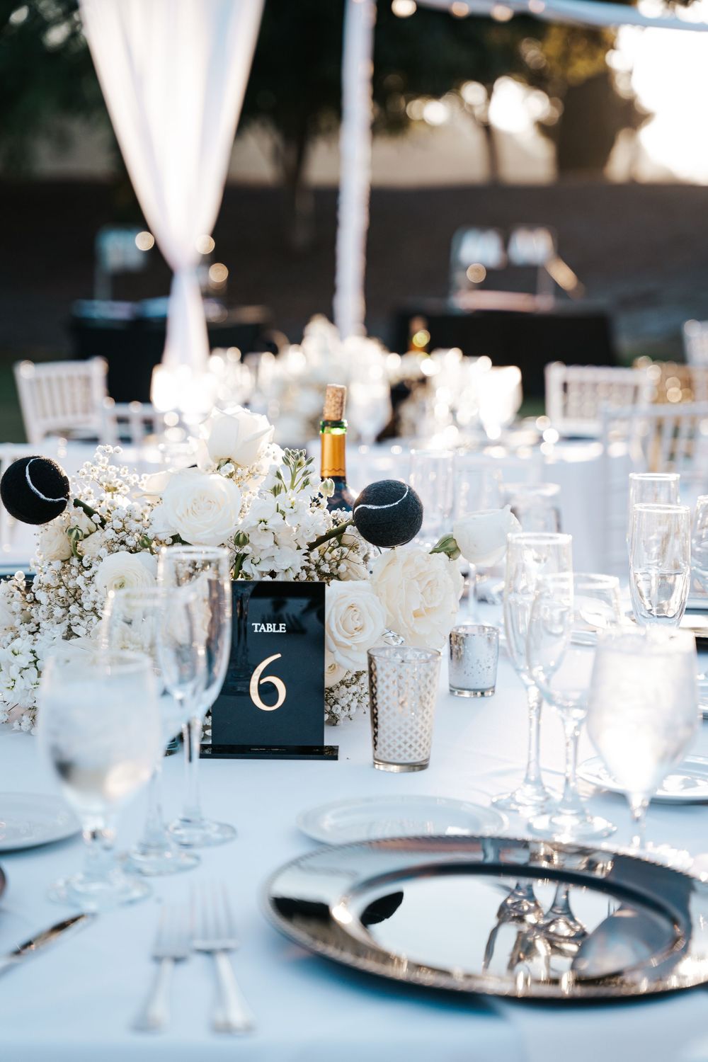 Elegant wedding reception tables with white floral centerpieces, crystal glasses, and black table numbers under outdoor tent.