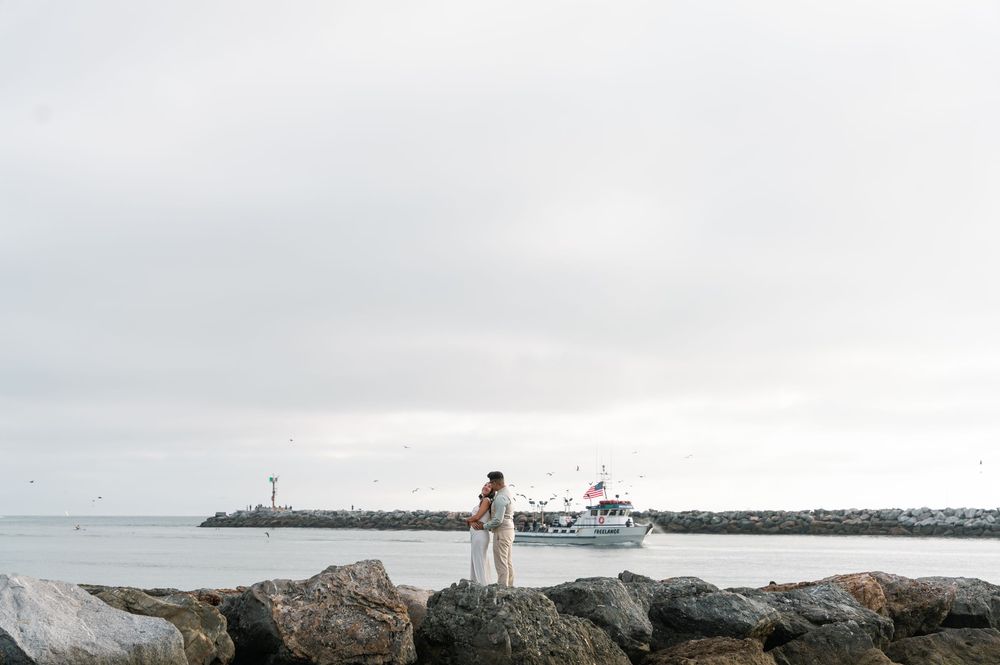 A panoramic view of a rocky coastal shoreline with a lighthouse and boat in the distance.