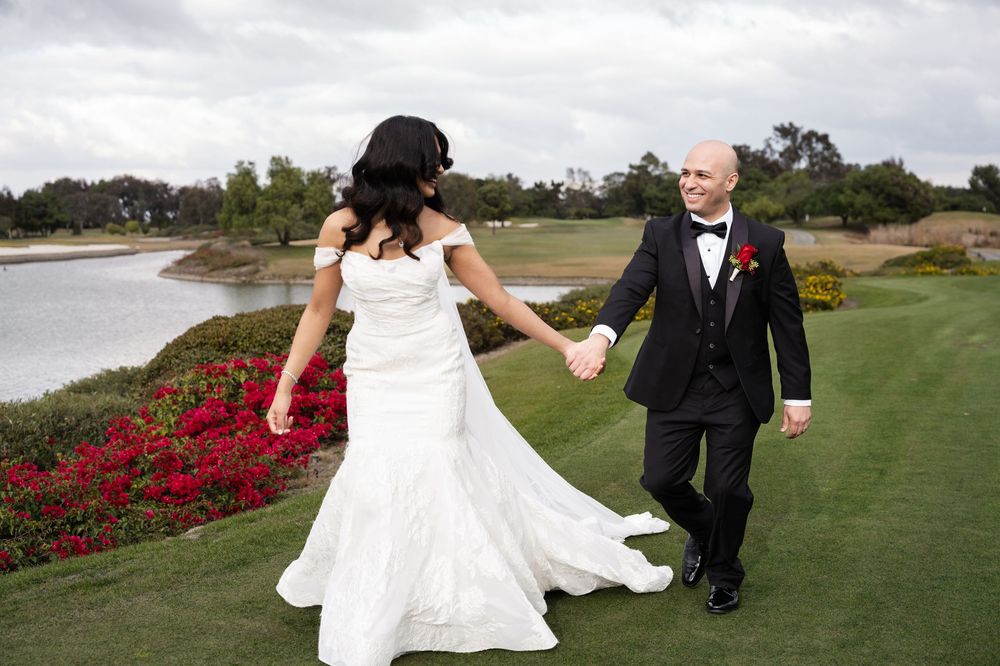 A couple in wedding attire holds hands while walking on a lush golf course with vibrant red flowers in the background.