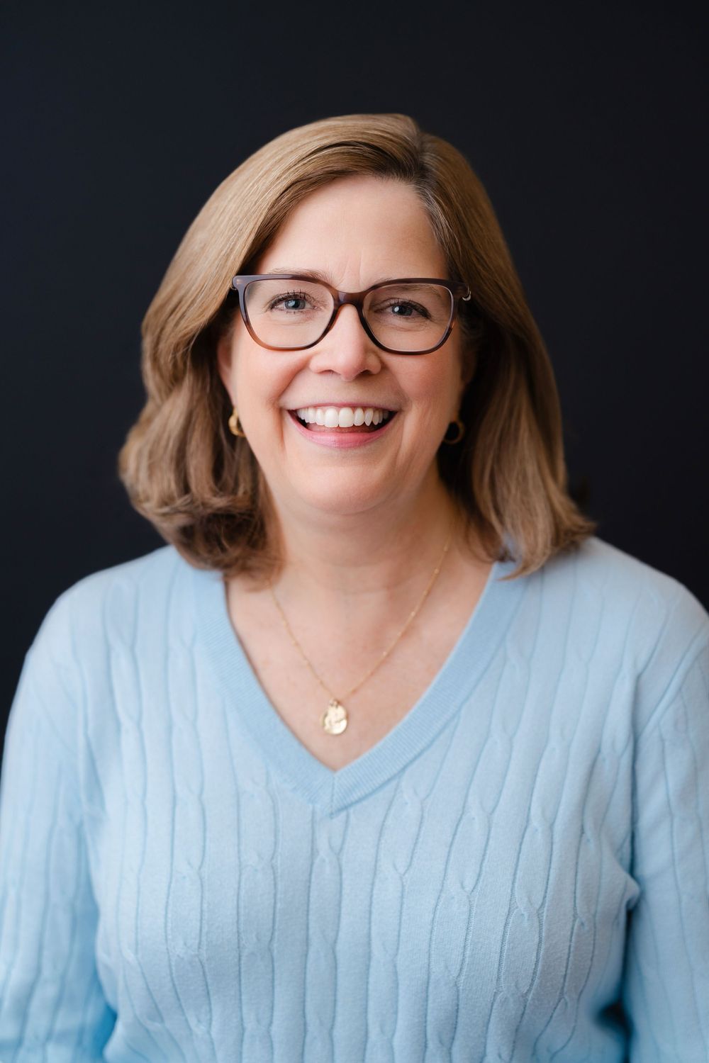Professional headshot of person in light blue sweater and glasses against dark background.