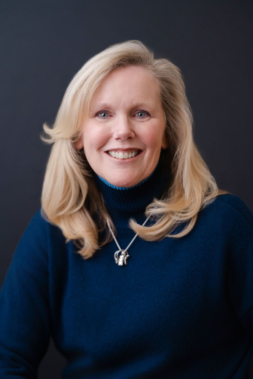 Professional headshot of a blonde woman in navy turtleneck sweater with silver necklace against dark background.