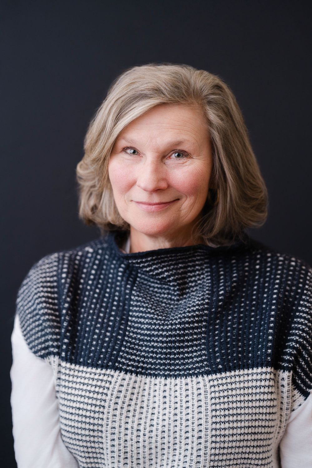 Professional portrait of a mature woman wearing a black and white patterned sweater against dark background.