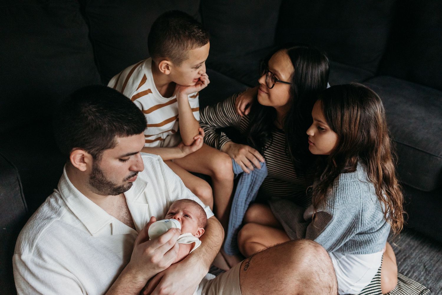 A family sits together on a dark couch, sharing an intimate moment while looking at their newborn baby.