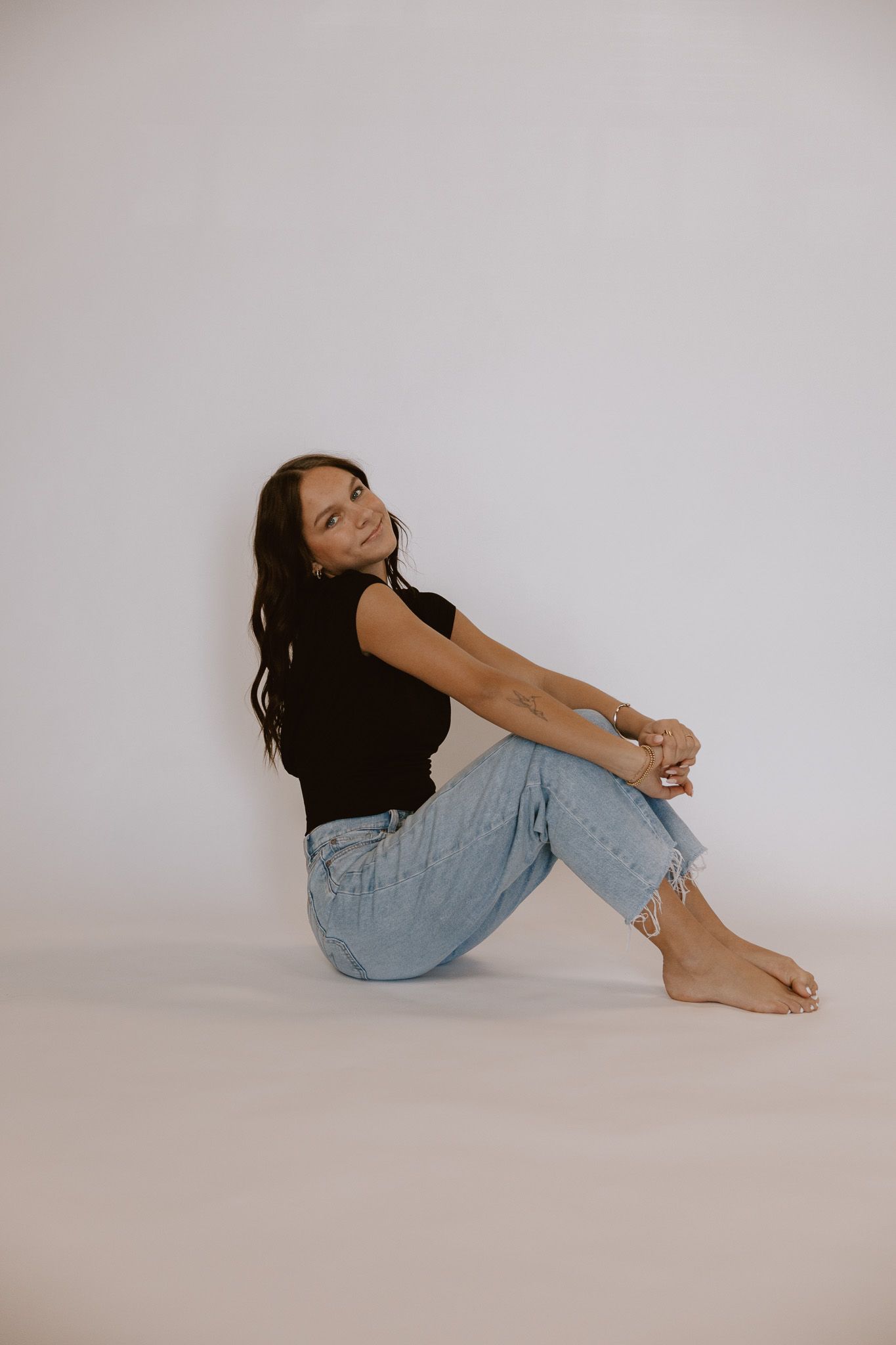 Young woman in black sleeveless top and ripped light jeans sitting barefoot on white studio floor.