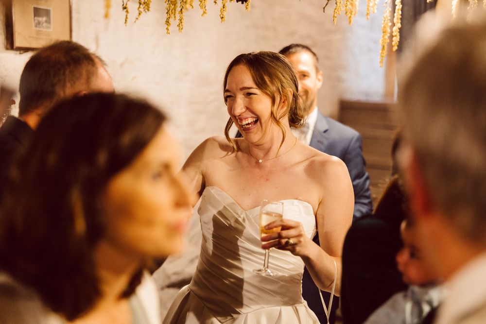 A joyful bride in a strapless white dress laughs while holding a drink at a wedding reception.