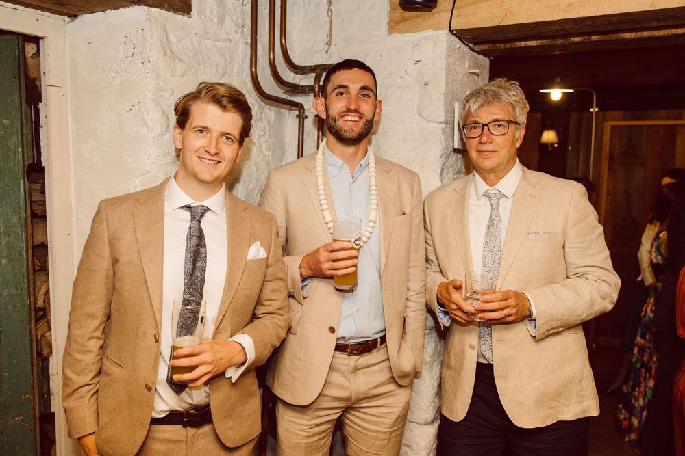 Three men in beige suits stand together holding drinks at an indoor event with rustic decor.