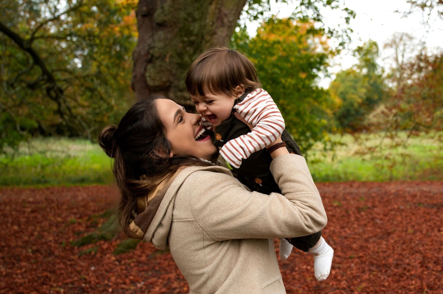 A parent holds and spins with a small child in a park filled with fallen autumn leaves on the ground.