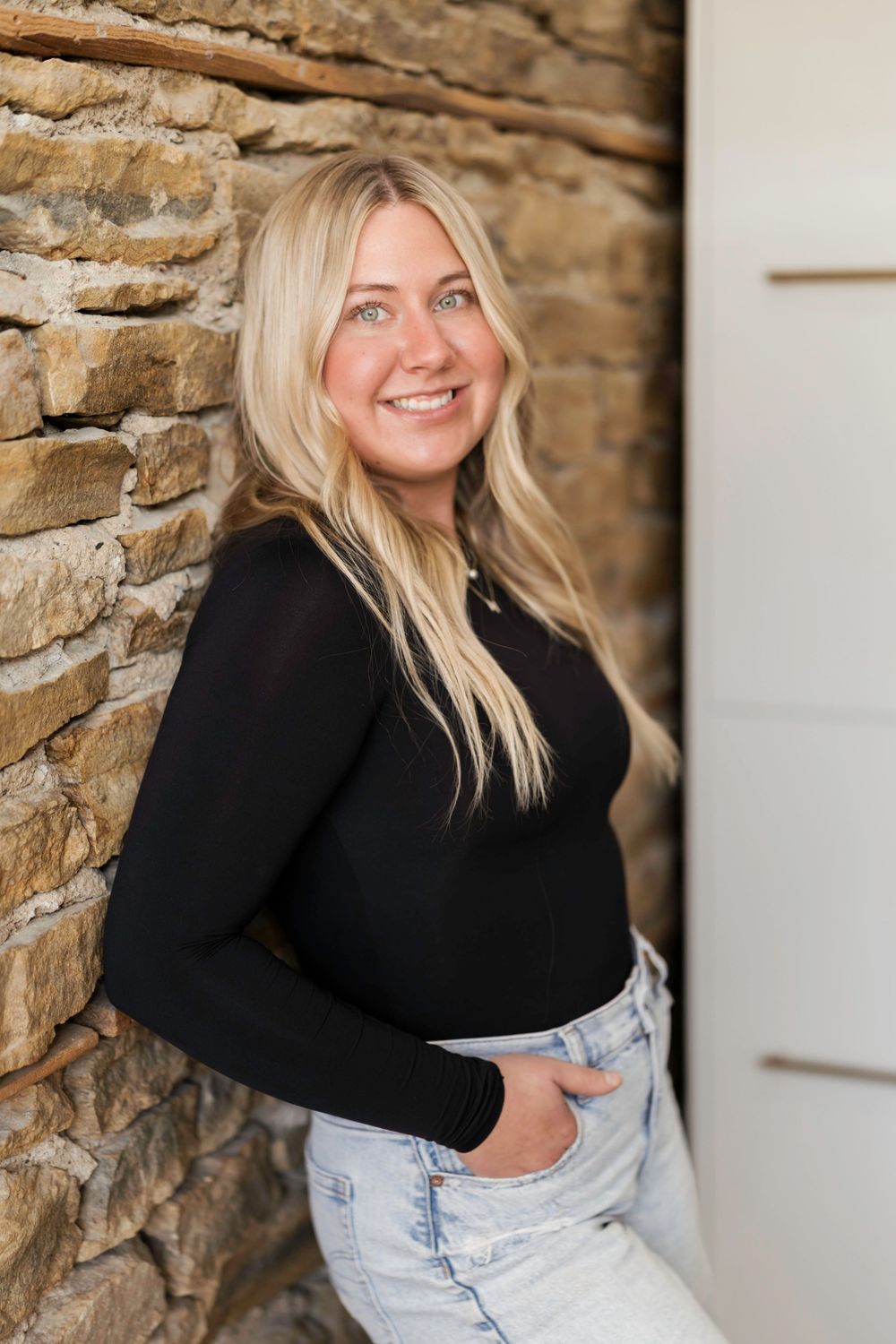 Person in black sweater and light wash jeans poses against rustic stone wall with natural lighting.