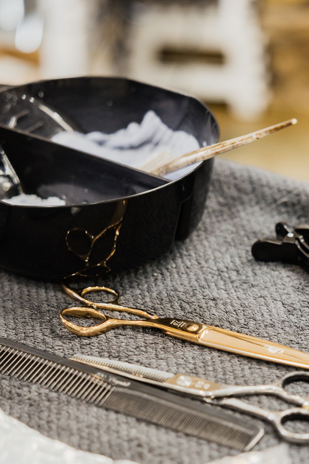 Professional hairdressing tools including gold scissors, combs and mixing bowl arranged on gray textured surface.