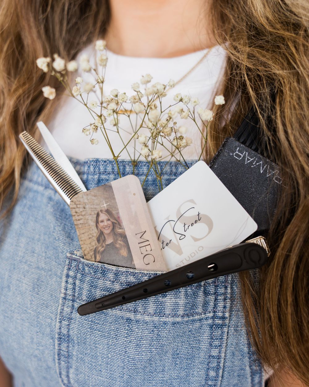 Hair accessories and dried flowers arranged artistically in denim overall pocket against light background.