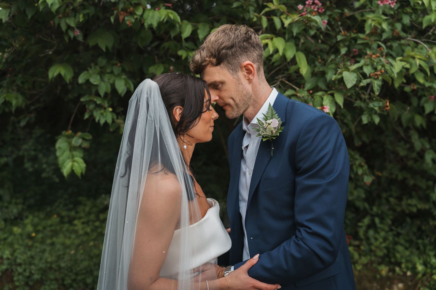 Bride and groom share an intimate moment together in a garden setting with lush greenery in the background.