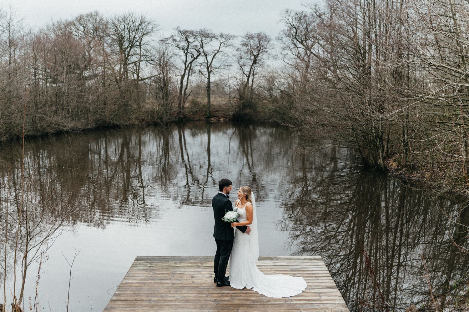 Winter wedding photo session on a wooden dock overlooking a serene lake surrounded by bare trees.