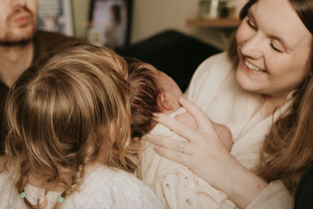 A smiling mother holds her newborn baby while an older sibling leans in to meet the infant for the first time.