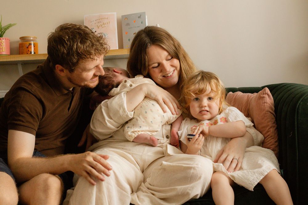 A young family of four sits together on a couch, with parents lovingly holding their newborn baby and toddler daughter.