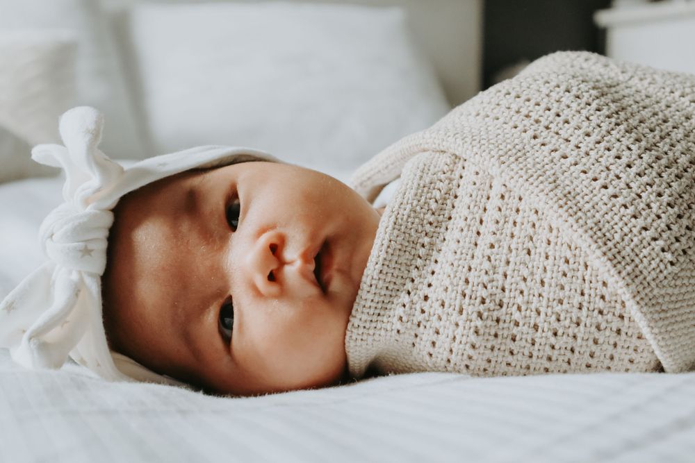 Baby wrapped in knitted blanket lying on white bedding with bright expression.