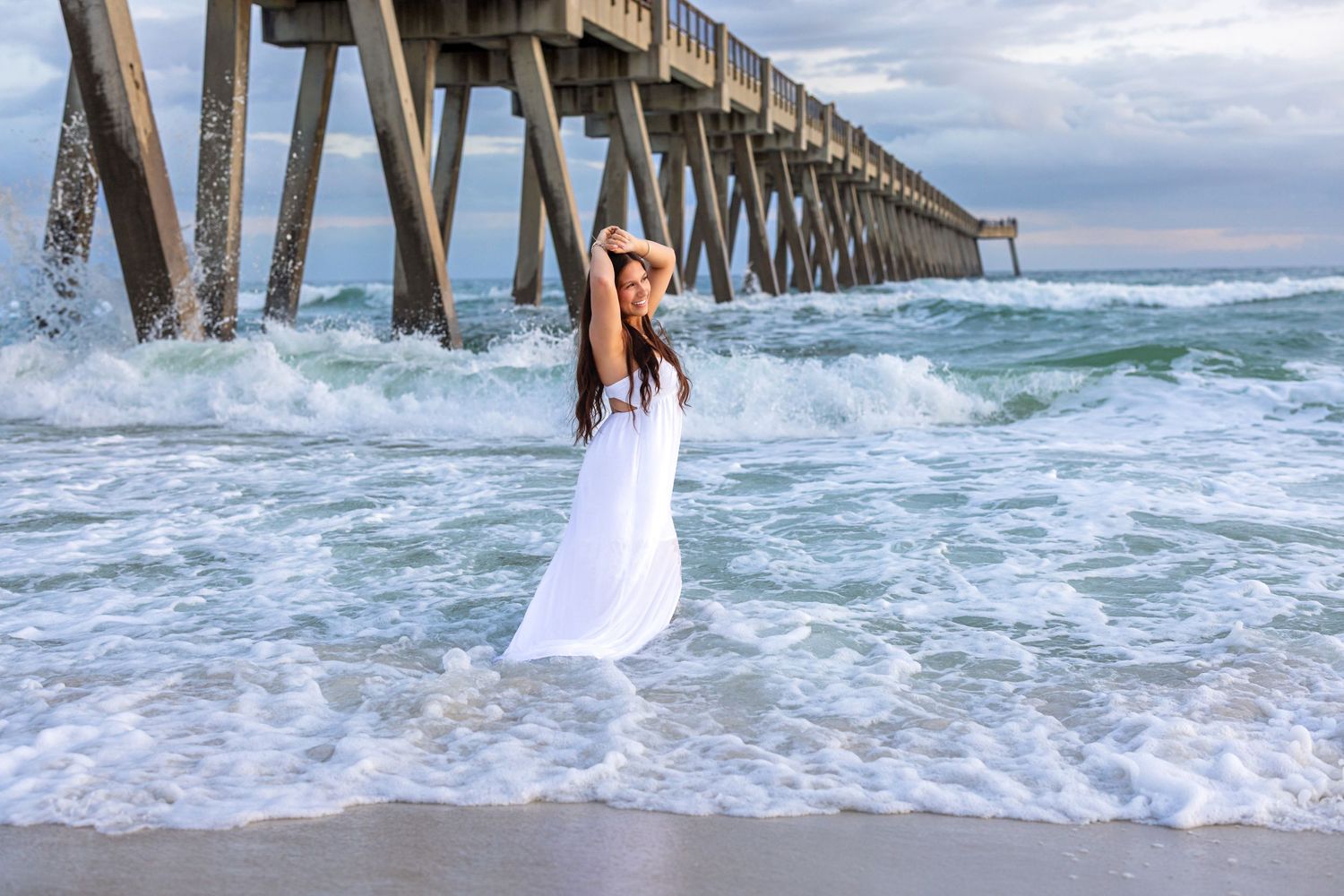 A person in a white dress walks along the shoreline near a wooden pier as waves crash against the beach.