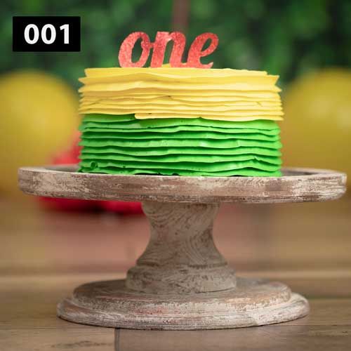 A layered birthday cake with yellow and green frosting and red 'one' topper on rustic stand.