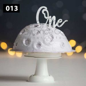 White frosted cake with elegant 'One' topper on display against dark background.