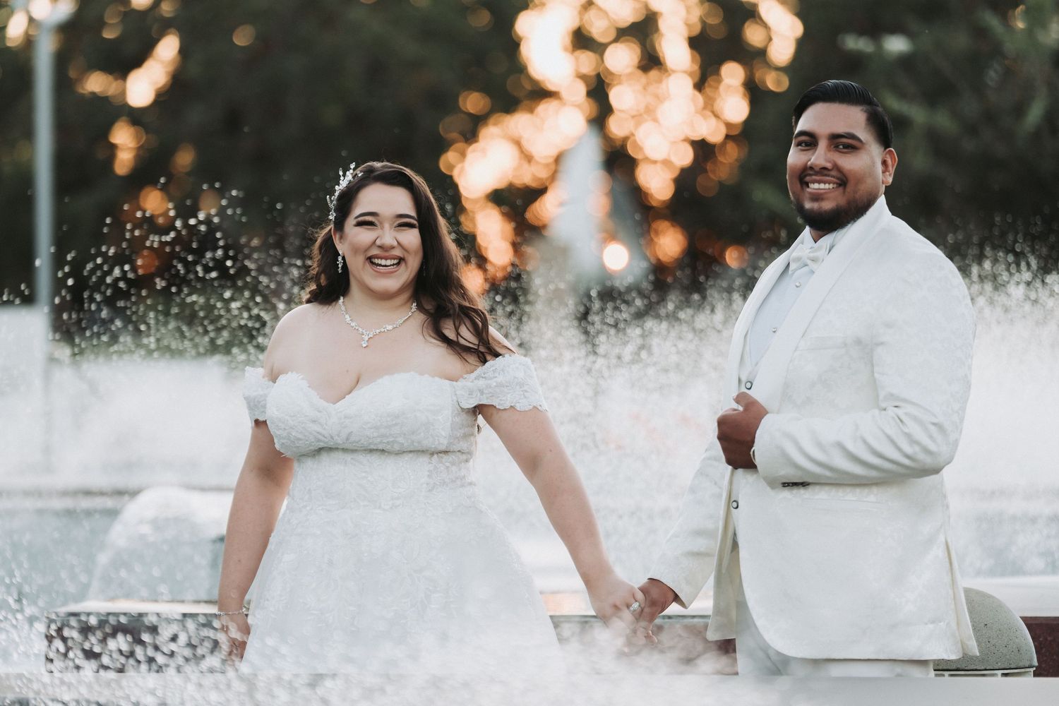 A couple in white wedding attire poses together outdoors at sunset with water fountain spray in the background.
