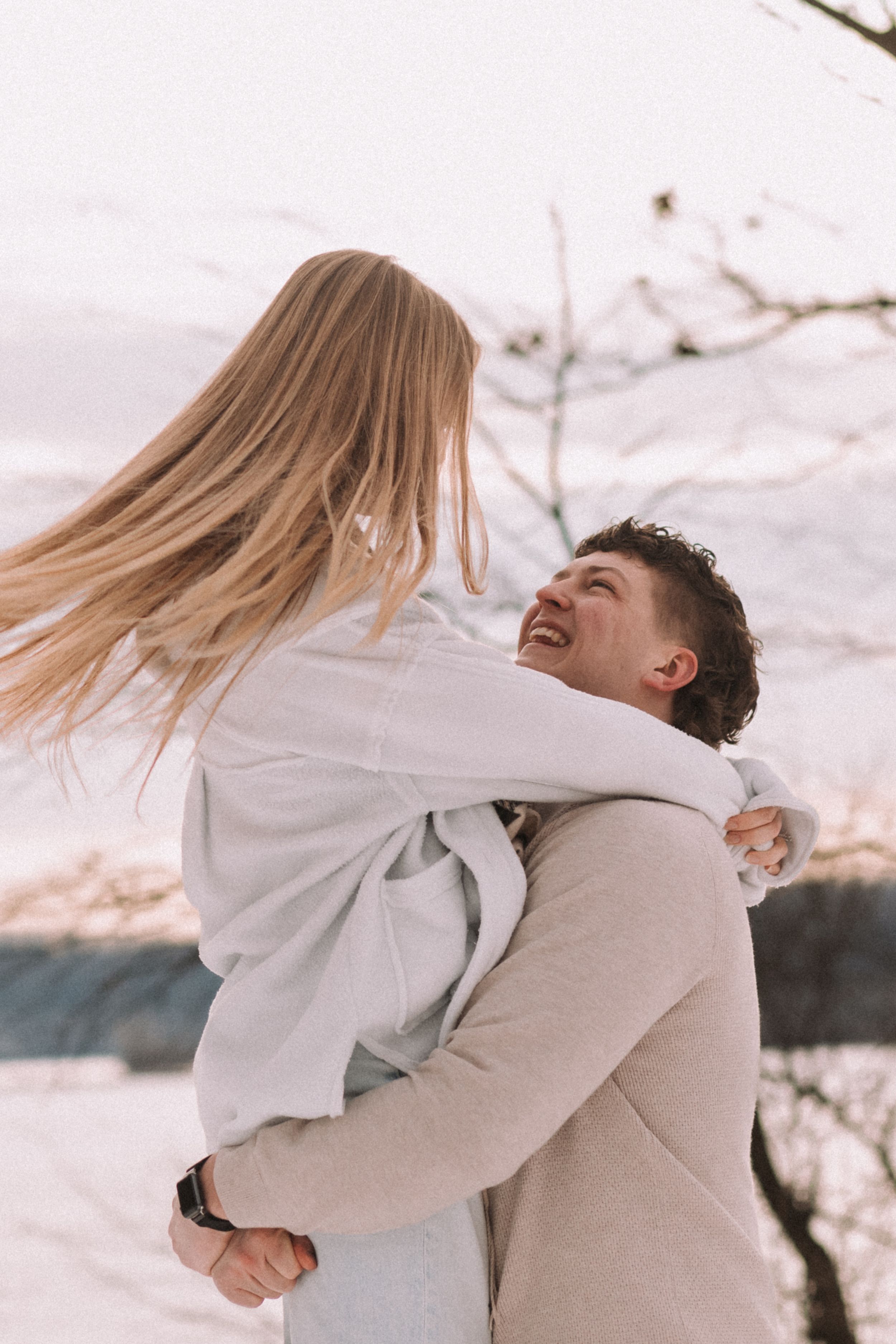 A couple embraces joyfully in a winter landscape with snow-covered trees in the background.