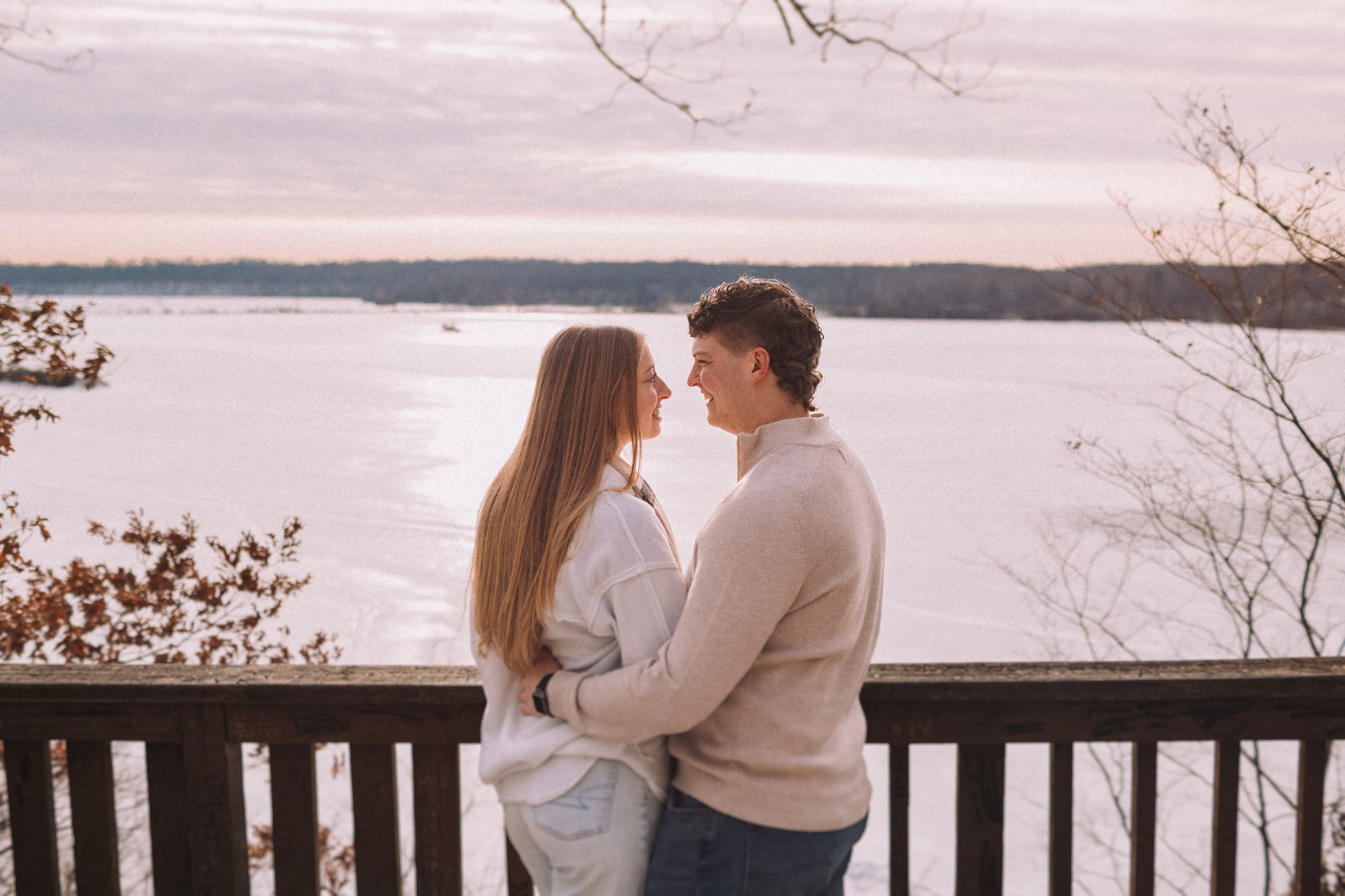 A couple in cozy sweaters embraces while looking out over a scenic lake view on an autumn day.