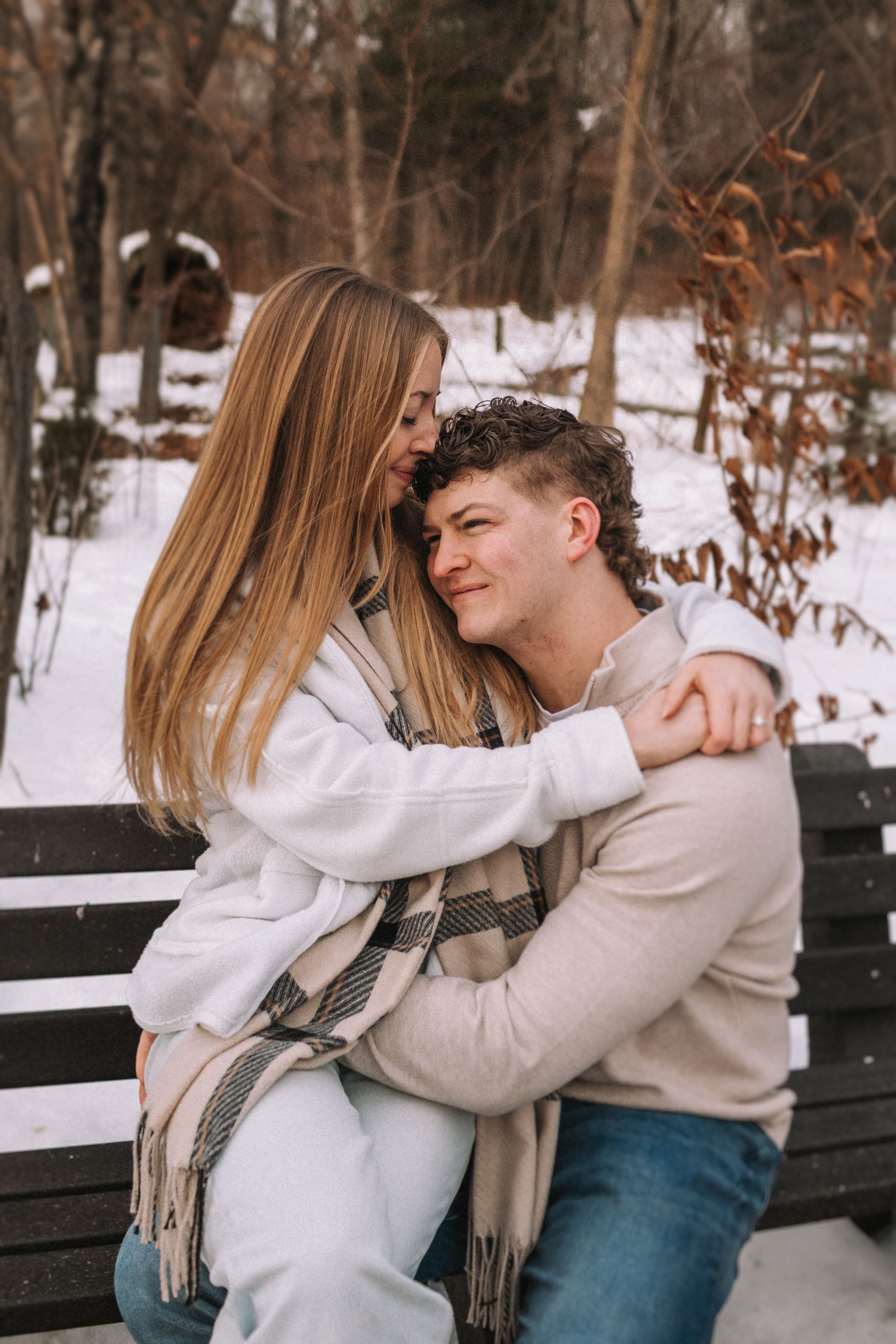 A couple embraces on a wooden bench in a snowy winter setting wearing light colored sweaters and scarves.