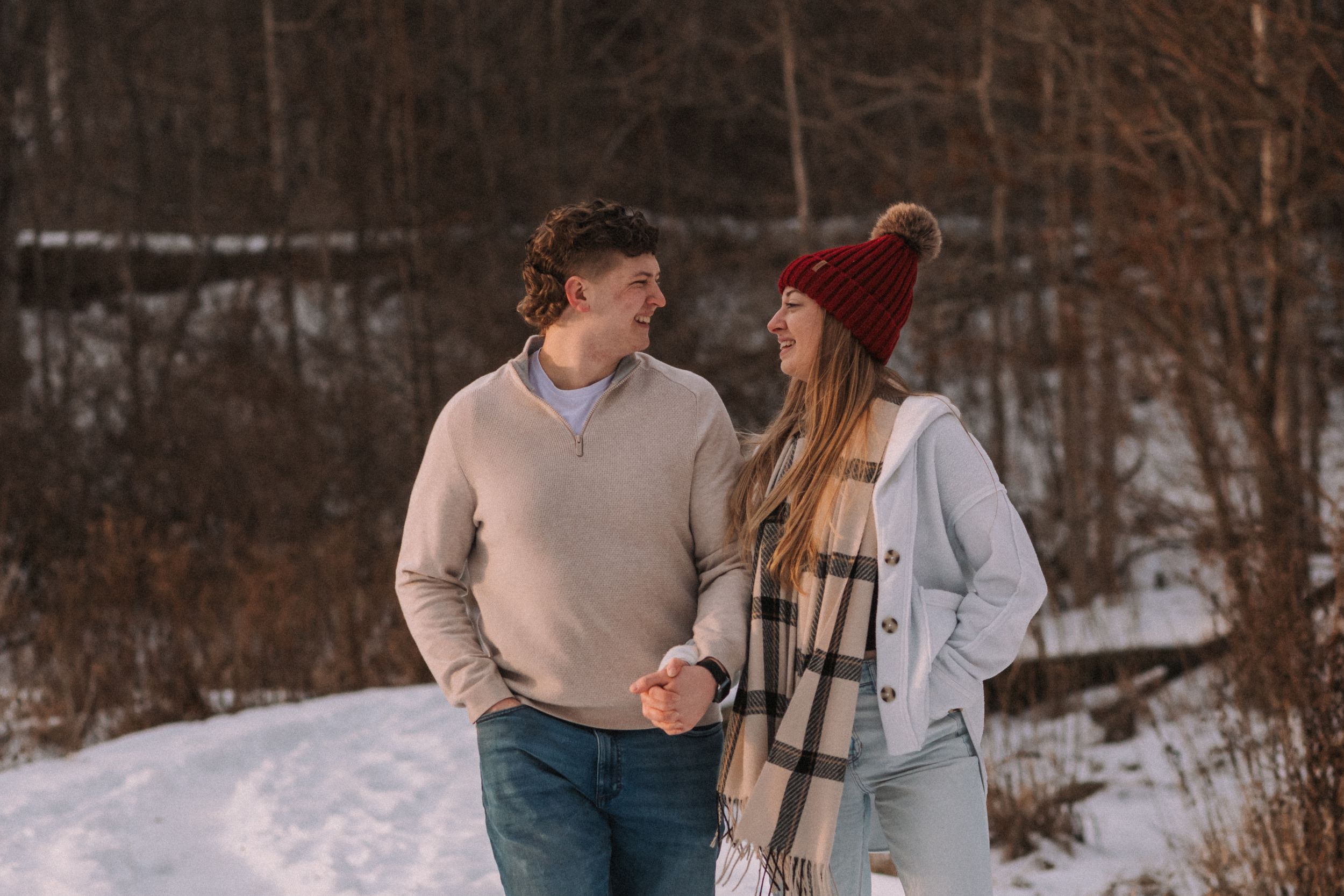 A couple holding hands while walking in a snowy winter landscape with trees in the background.