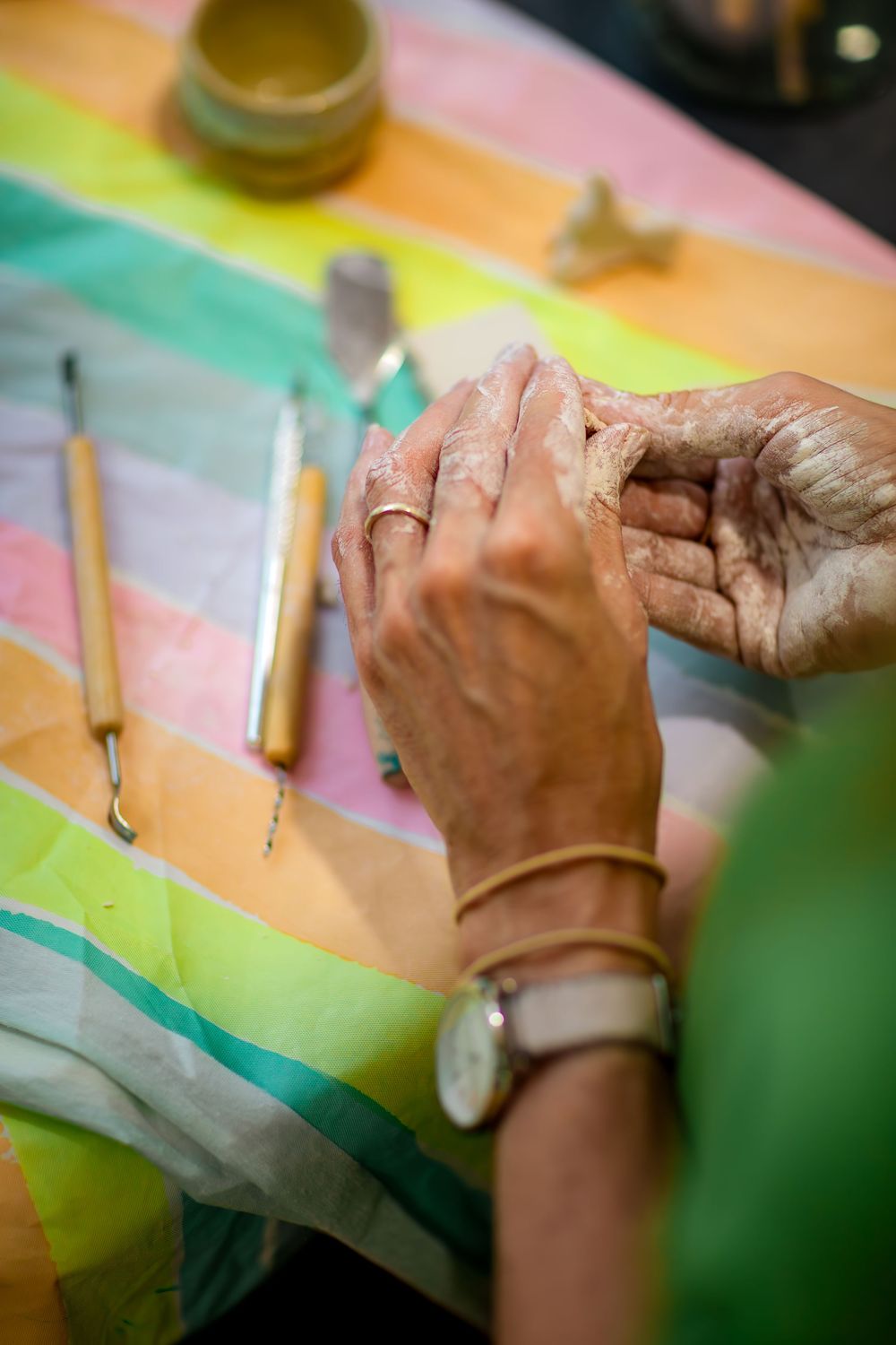 Paint-stained hands work on a colorful rainbow striped artwork with brushes and art supplies on the table.