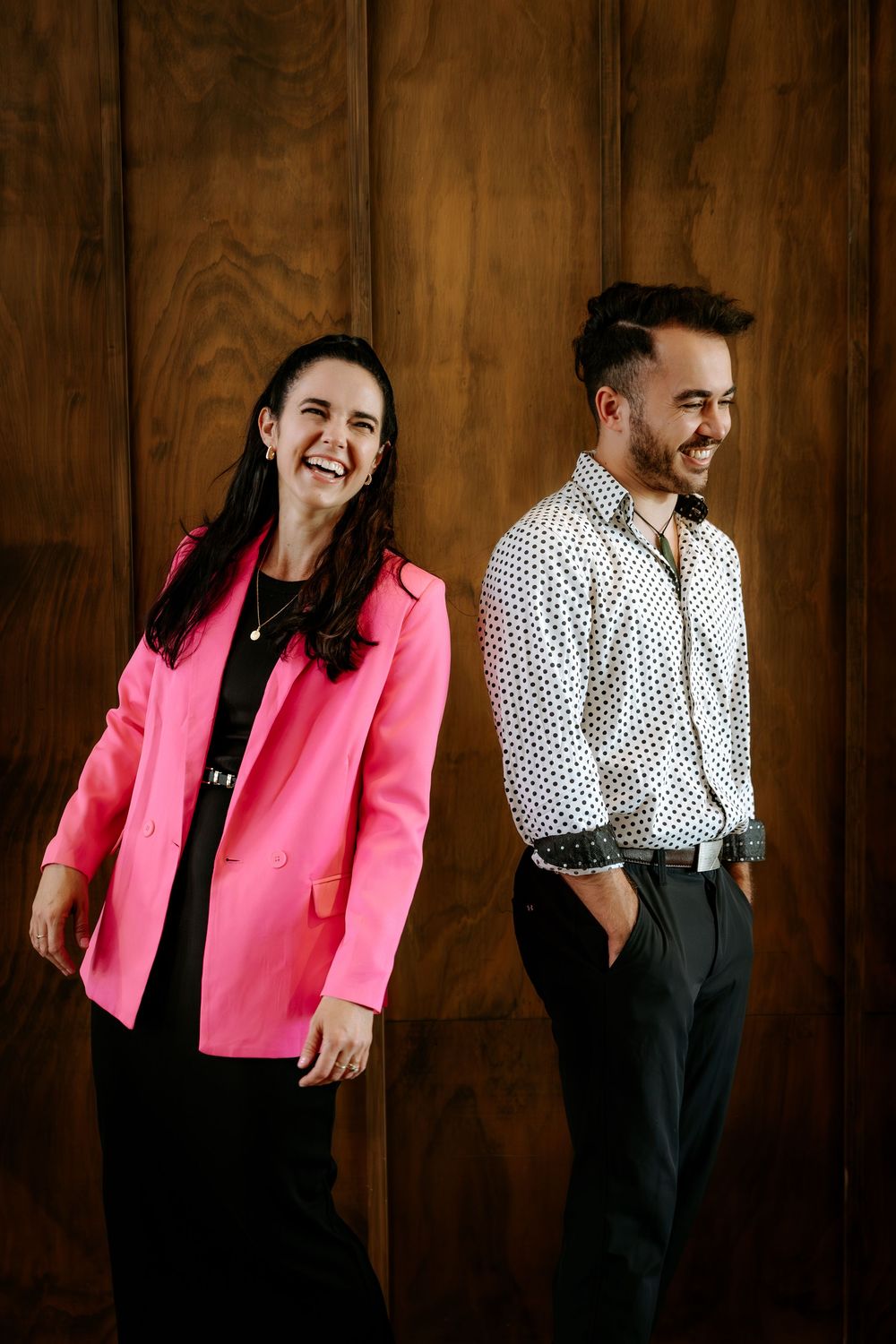 A couple in pink blazer and polka dot shirt share candid laughs against a wooden wall in a photo series.