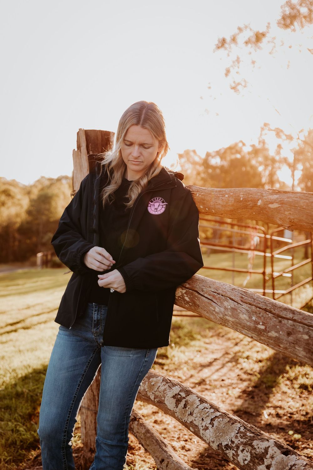 Woman in black jacket and jeans leaning on rustic wooden fence at golden hour on a farm.