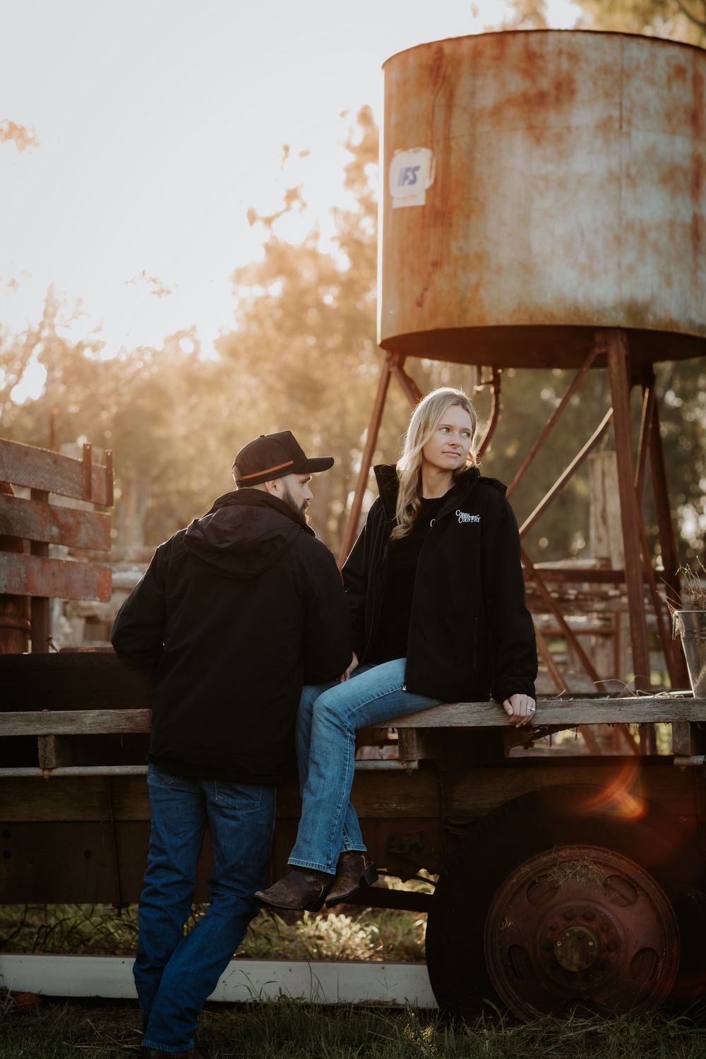 A man in a cowboy hat talks to a blonde woman sitting on a rustic farm trailer at golden hour.