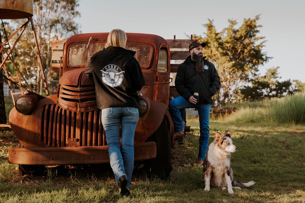 Two people and a dog stand beside a rustic vintage red truck on a farm during golden hour.